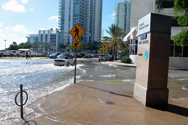 A road on left, covered in sea water, is being driven through by a car. The sun is shining indicating that the water did not fall as rain but is instead due to the tides.